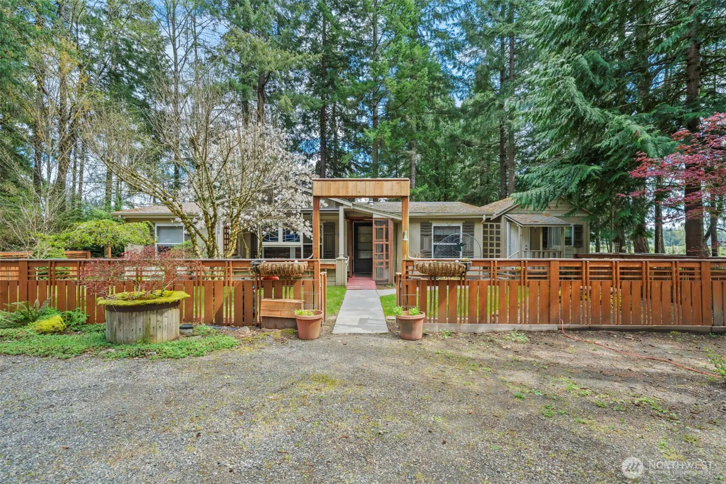 A tan house with a brown fence, a gravel driveway, and tall green trees in the background.