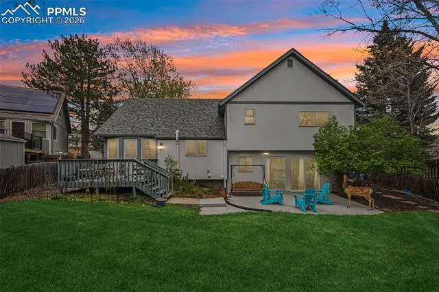 Backyard view of a two-story gray house with a deck, patio, and green lawn at sunset. Blue chairs and a swing sit on the patio. A deer stands near a tree.