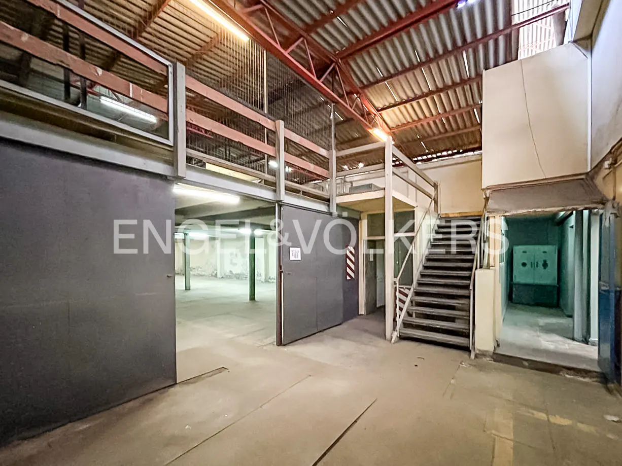 Interior view of a large, empty warehouse with a metal roof and a second-story platform with stairs.