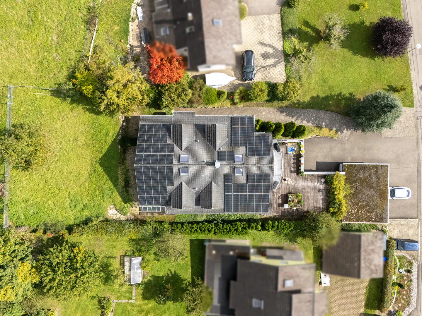 Aerial view of a house with extensive solar panels on its roof, surrounded by a green lawn, trees, and a paved driveway.