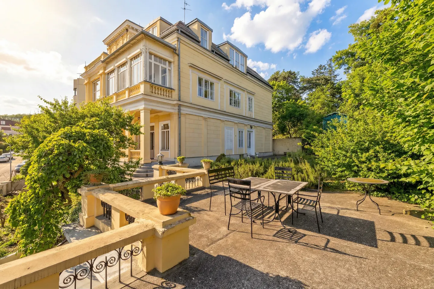 Exterior view of a yellow, multi-story house with a patio featuring a table and chairs. Lush greenery surrounds the property.