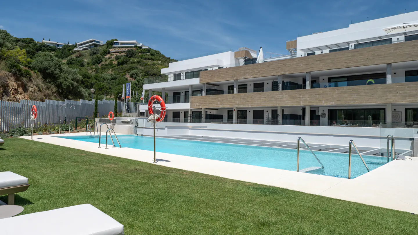 Outdoor pool with turquoise water, surrounded by green grass and white buildings under a blue sky. Life preservers are visible.