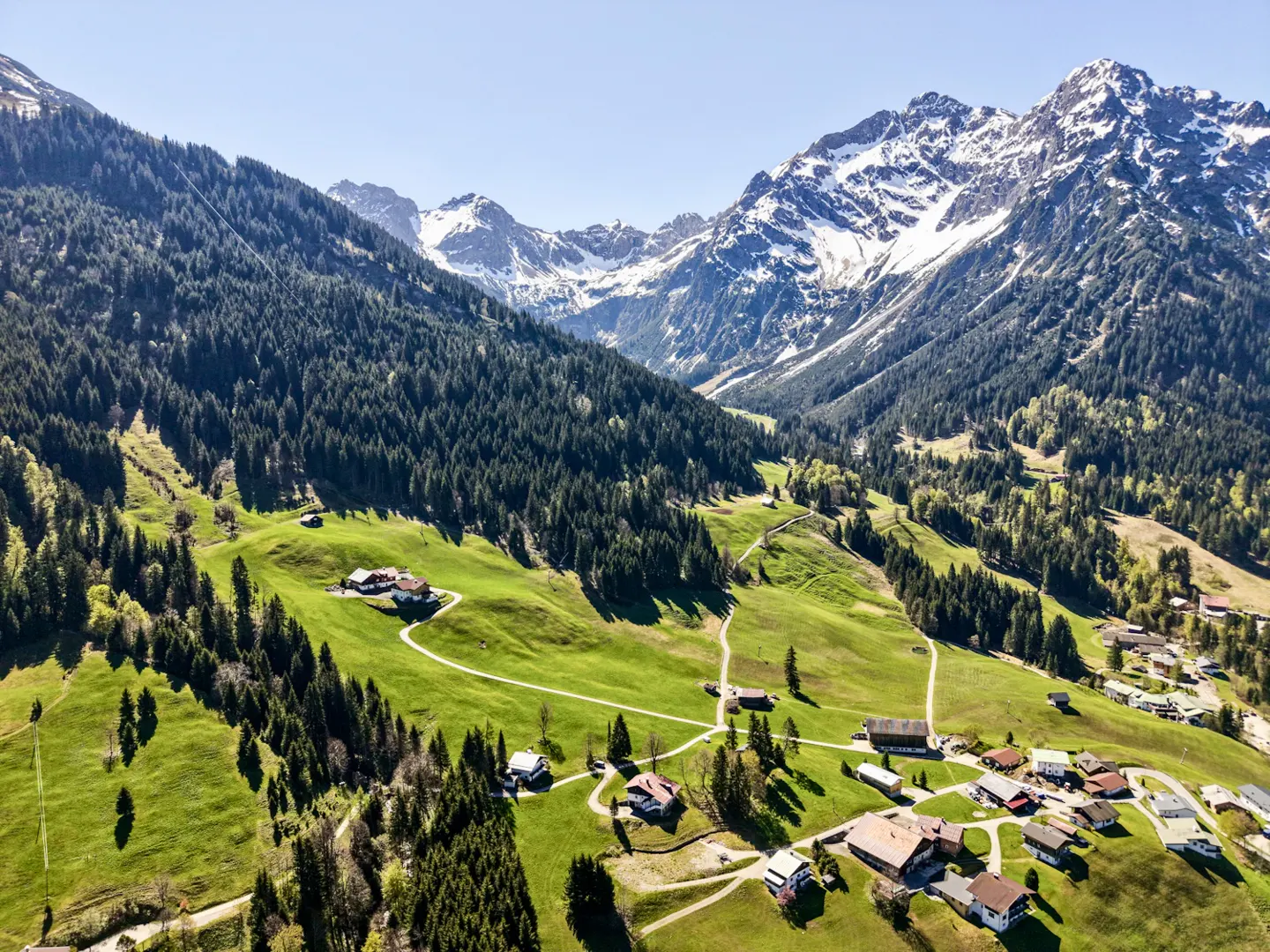 Scenic aerial view of a green valley with houses, surrounded by forested hills and snow-capped mountains under a clear blue sky.