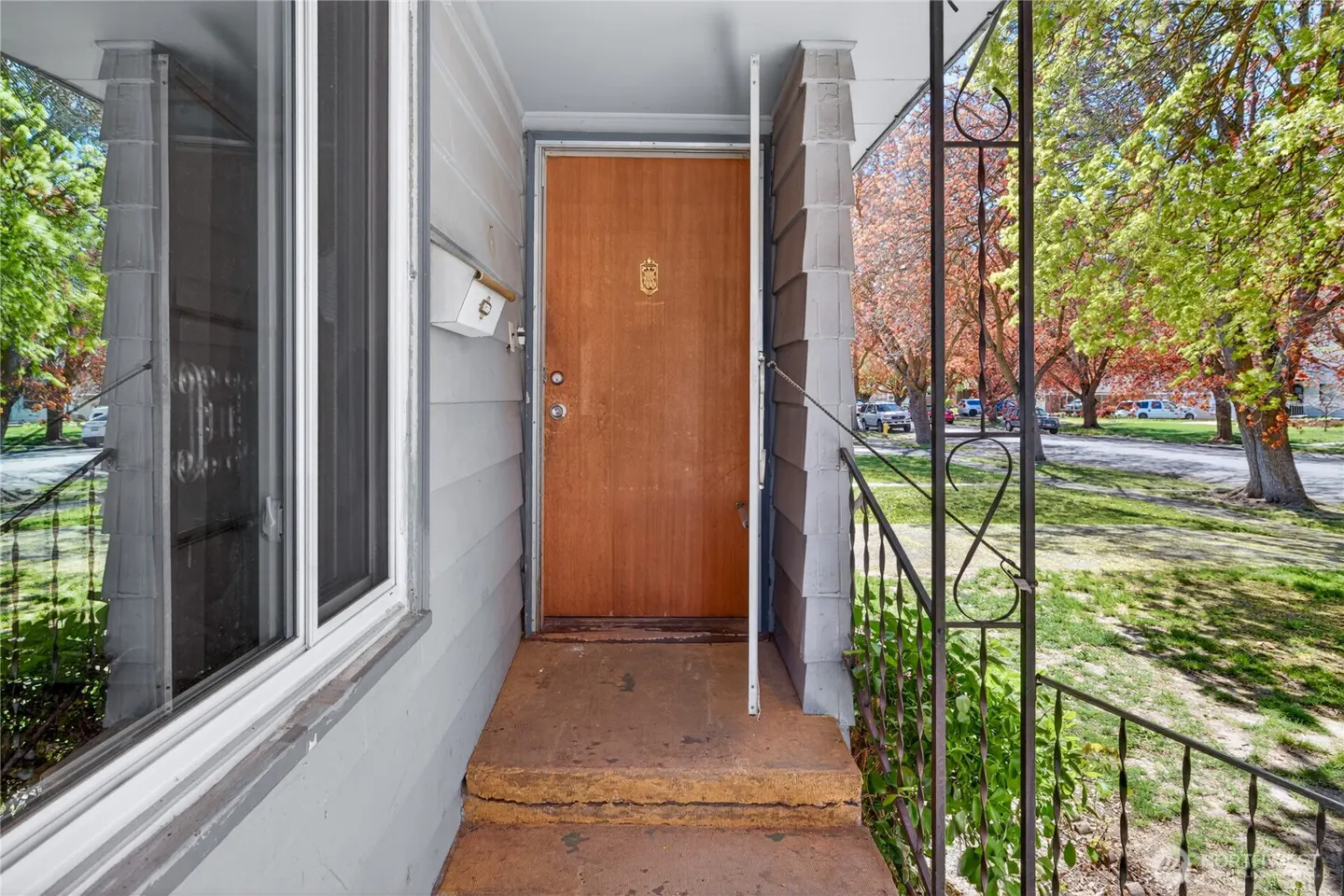 Front entrance of a gray house with a brown door, concrete steps, and a black iron railing. Trees and a street are visible in the background.