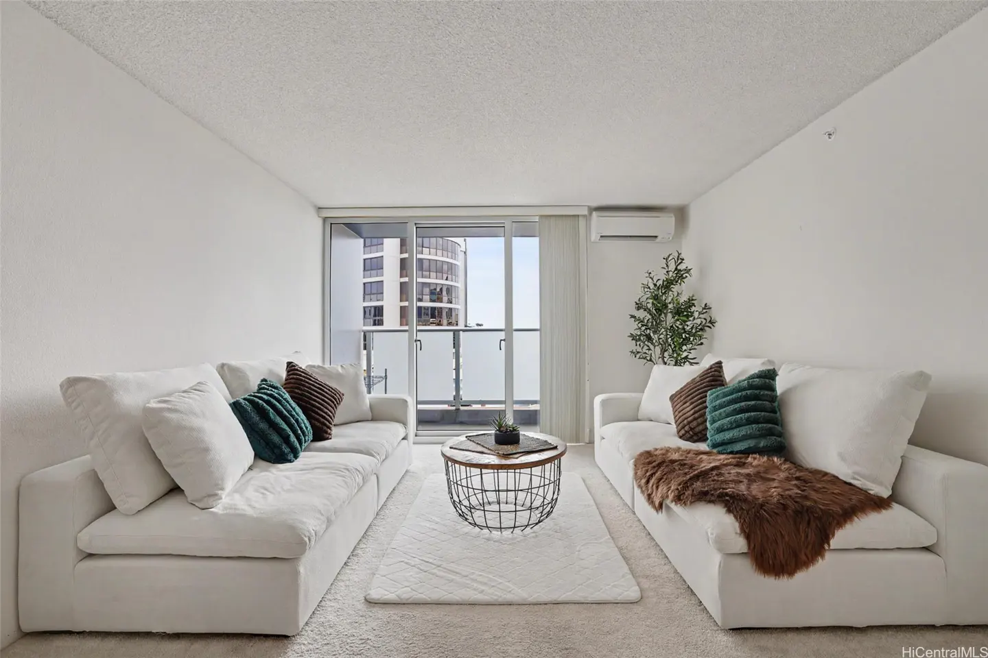 Bright living room with two white sofas facing each other, teal and brown pillows, a wire coffee table, and a balcony with a city view.