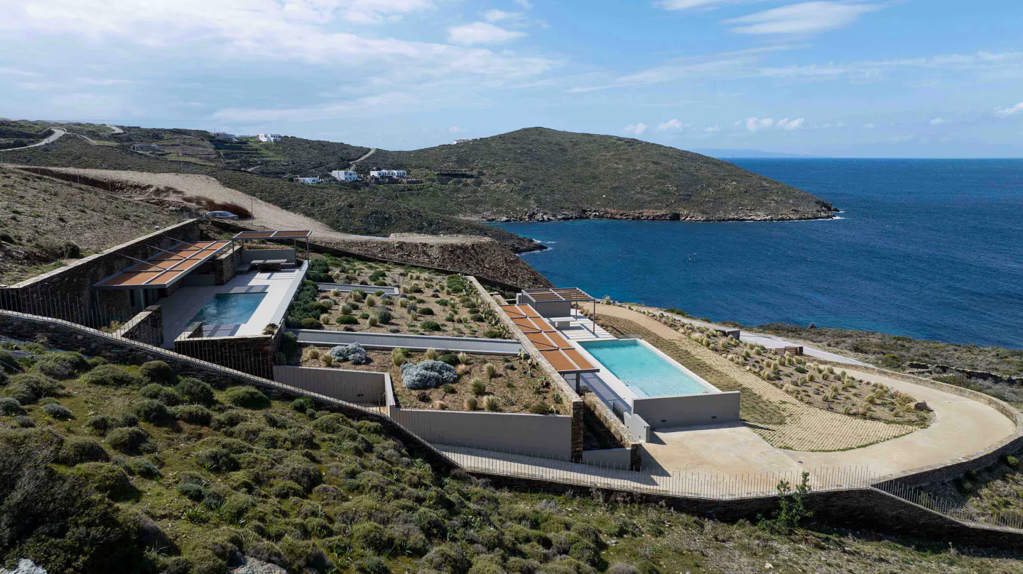 Luxury villa with pools on a hillside overlooking the blue ocean under a cloudy sky.