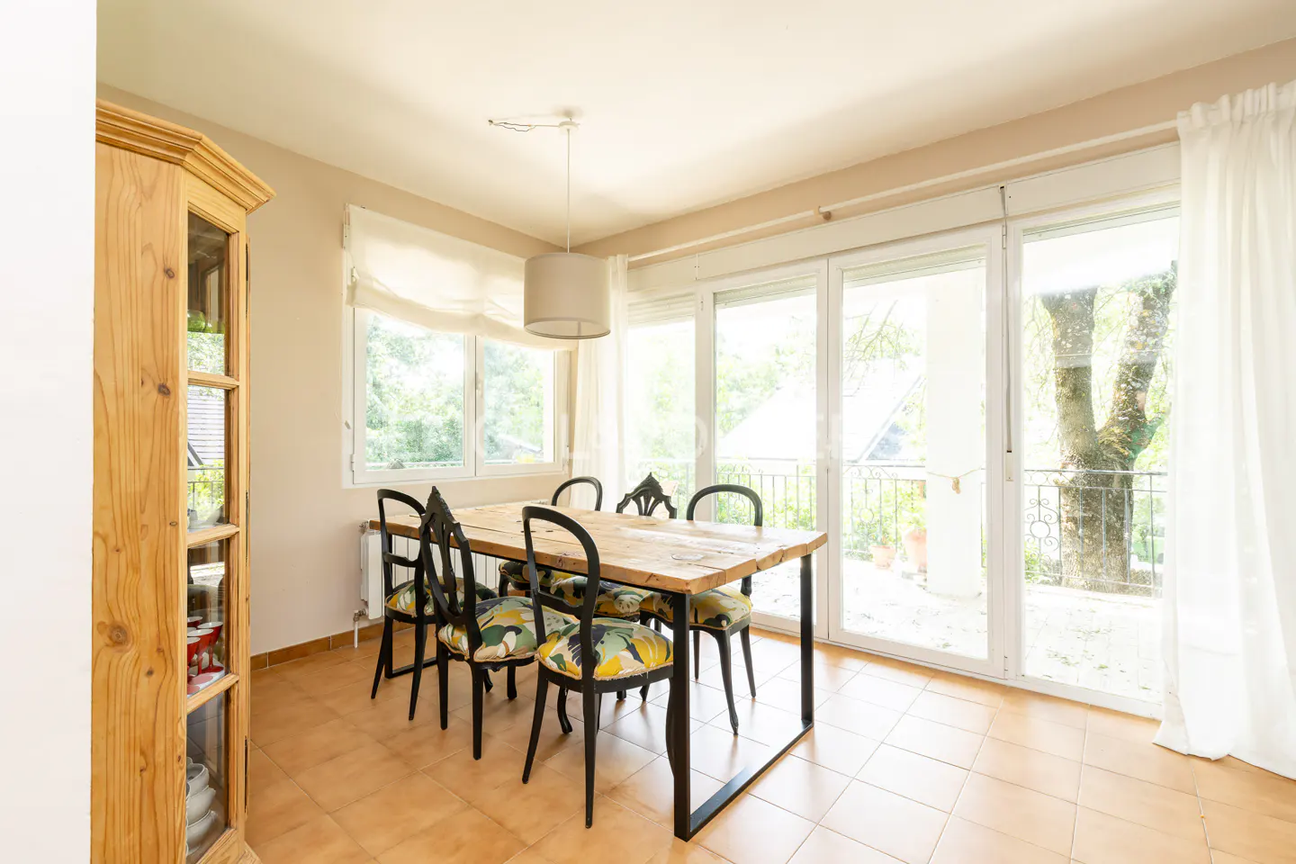 Bright dining room with a wooden table, black chairs with floral cushions, and a light fixture. Large windows overlook a balcony.