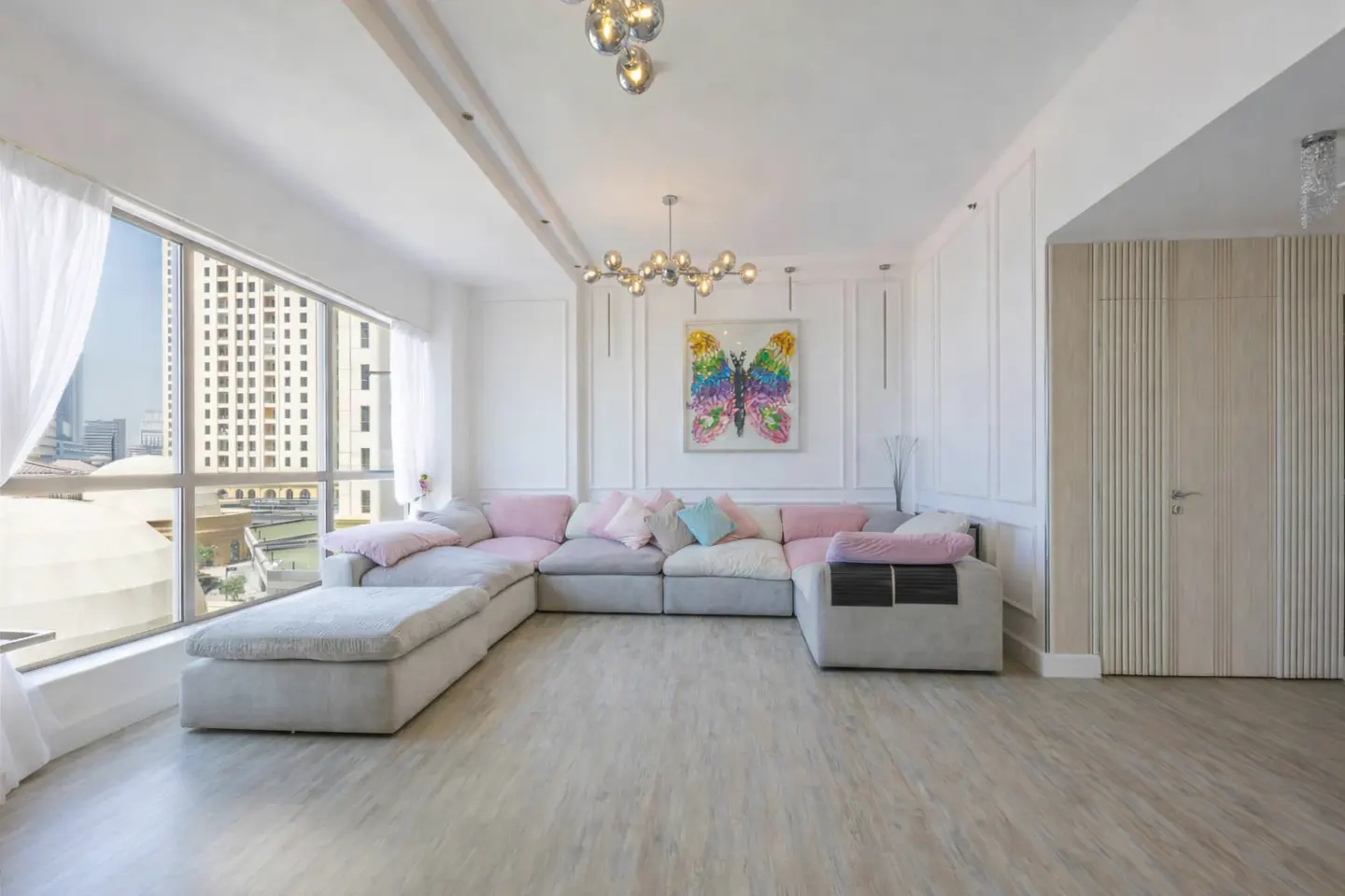 Bright living room with a large window, a gray sectional sofa with pink pillows, and a colorful butterfly painting on the wall.