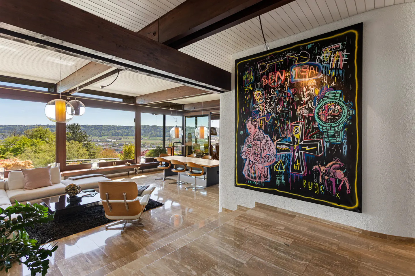 Open-plan living room with marble floors, exposed beams, and a large abstract painting on the wall. Outside, there is a view of trees and a town.