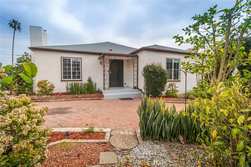 Front view of a white stucco house with a gray roof, brick driveway, and lush landscaping.