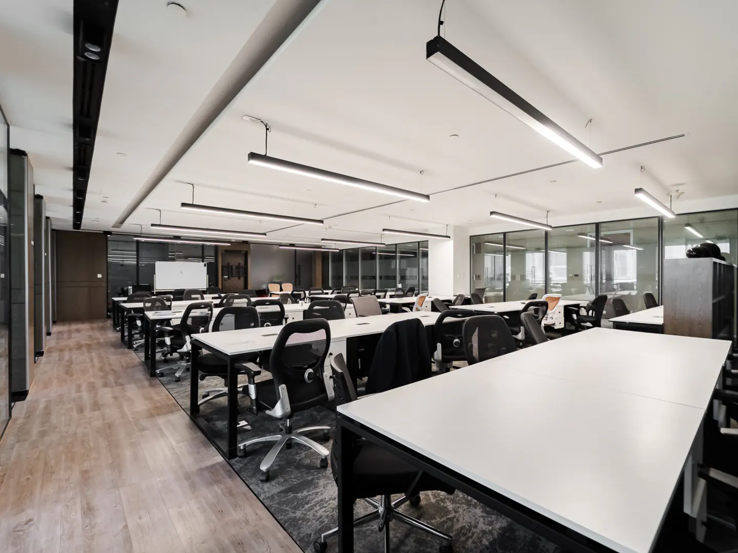 Bright, modern office space with rows of white desks and black chairs. Glass-walled offices line the perimeter. Linear lighting overhead.