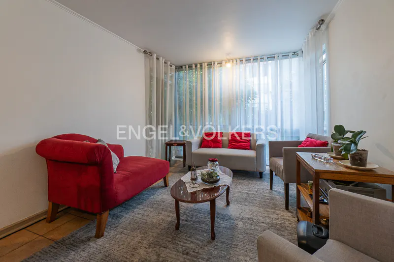 Living room with a red chaise lounge, gray sofa with red pillows, and a coffee table on a gray rug. White walls and sheer curtains.
