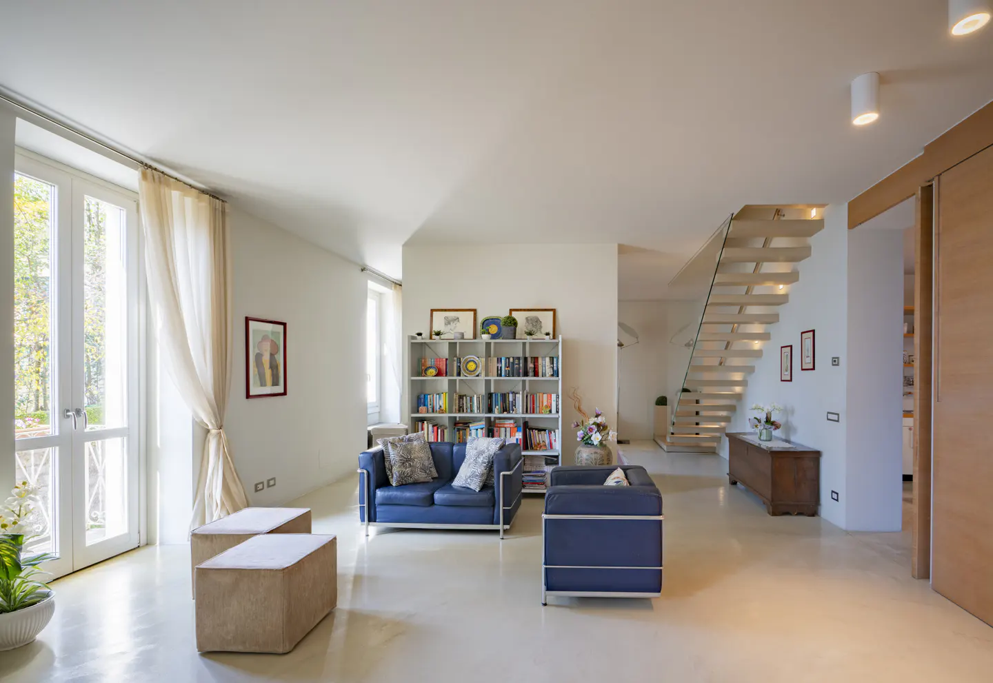 Bright, modern living room with white walls, a blue sofa, a white bookshelf, and a staircase with glass railings. Natural light streams through large windows.