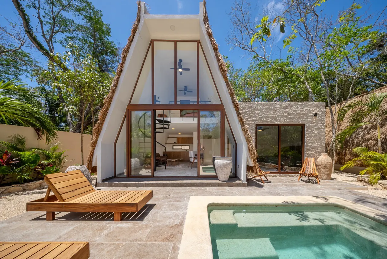 A-frame house with a thatched roof, a pool, and a wooden lounge chair on a stone patio.