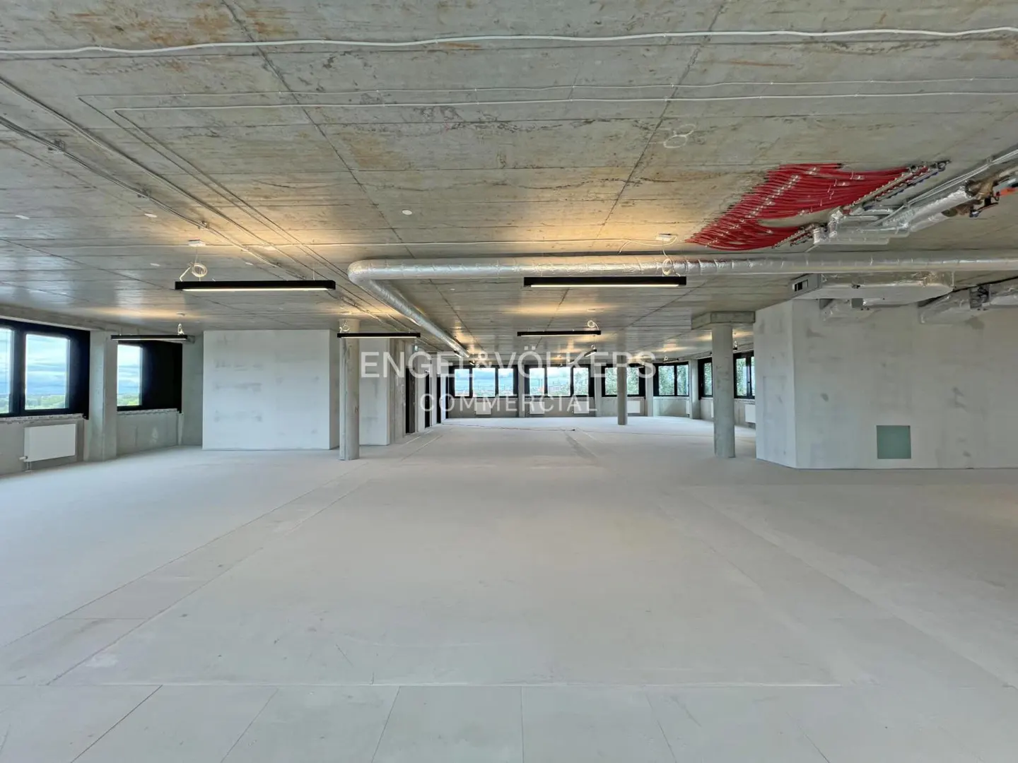An empty commercial space with concrete floors, pillars, and ceiling. Windows line the walls, and the Engel & Völkers logo is visible.