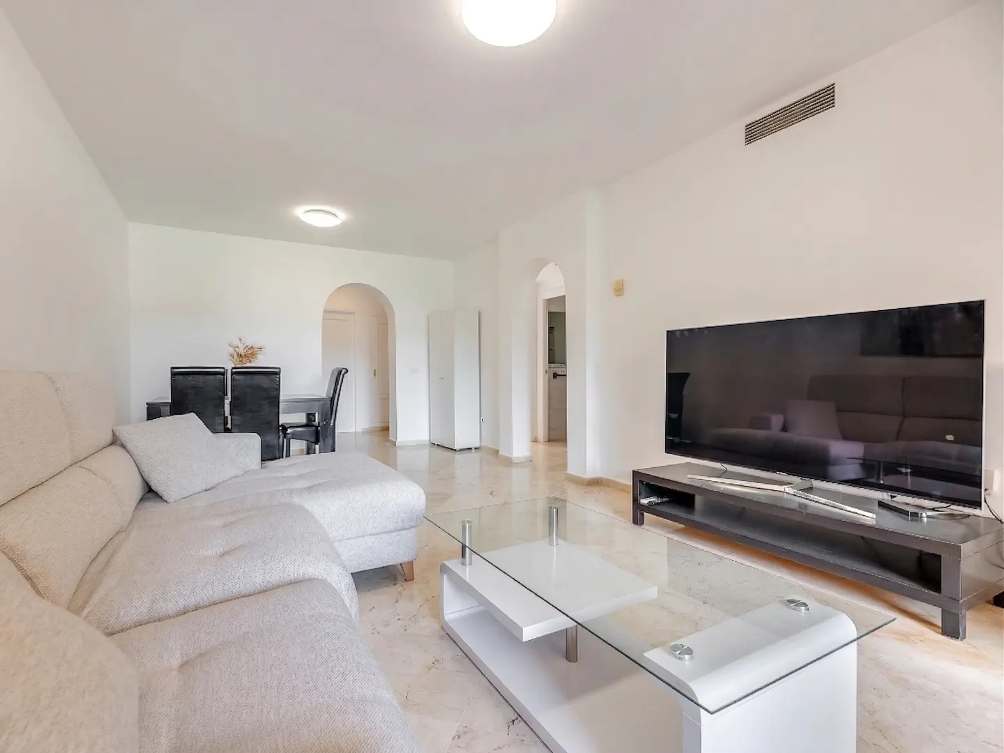 Bright living room with a beige sectional sofa, glass coffee table, and a large TV on a black stand. Dining table with black chairs in the background.