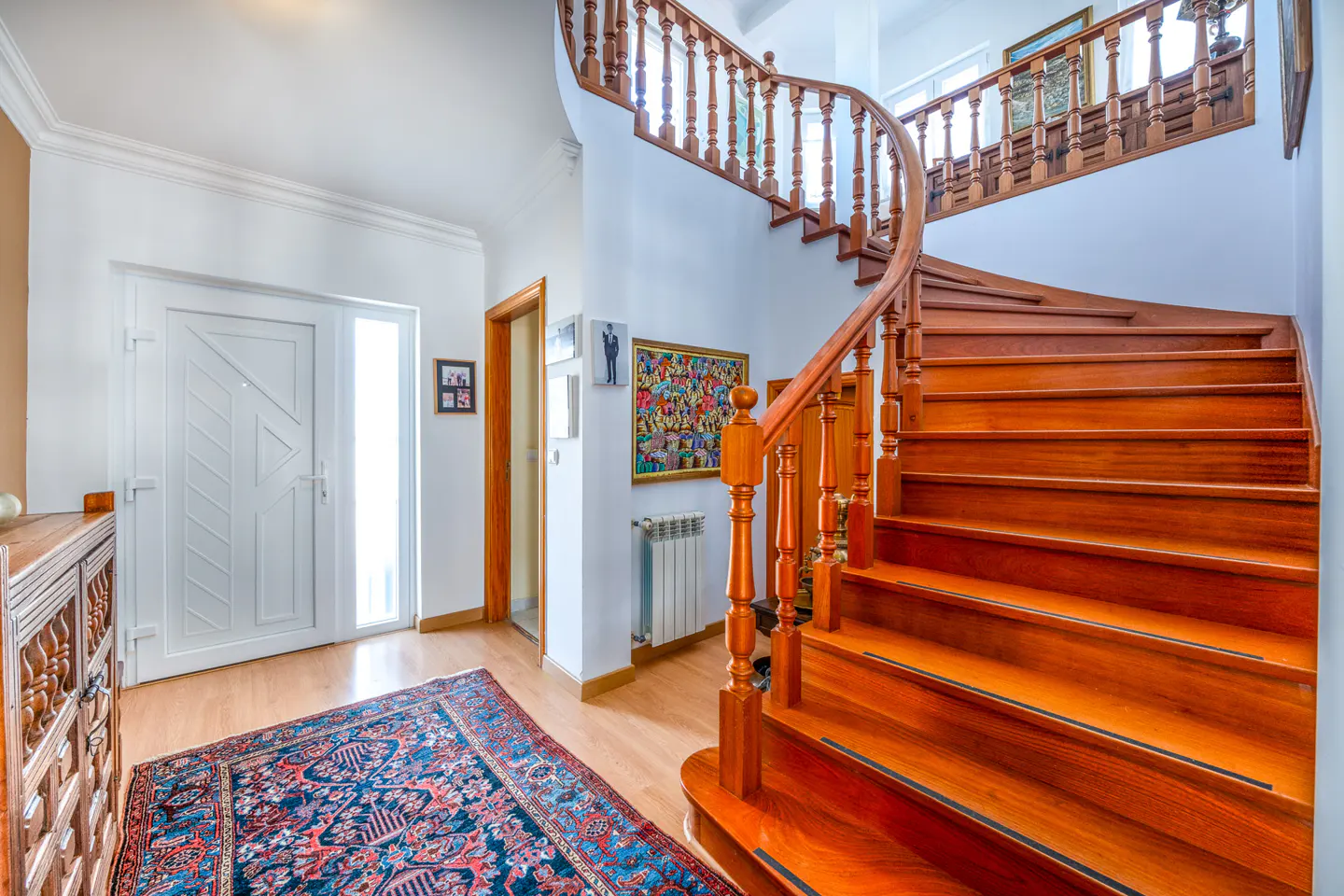 Bright foyer with a curved wooden staircase, white walls, and a blue patterned rug on a light wood floor. A white door is visible.