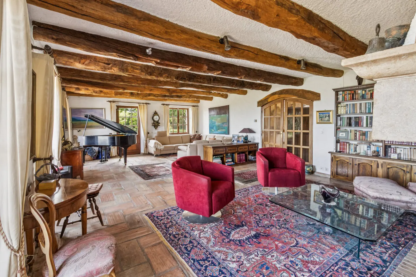 A spacious living room with exposed wooden beams, red armchairs, and a grand piano.