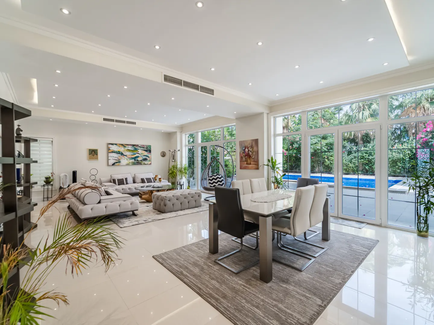 Bright, open-plan living and dining area with white walls, tile floors, and large windows overlooking a pool and garden.