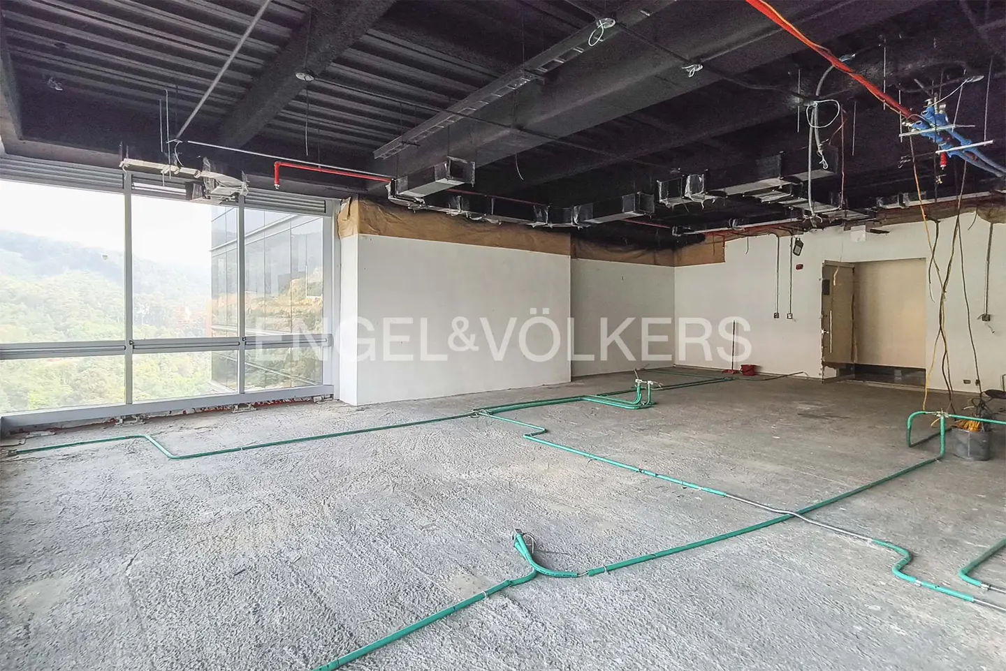 Unfinished office space with exposed ceiling, concrete floor, and green pipes. Large windows offer a view of greenery. "Engel & Völkers" logo visible.