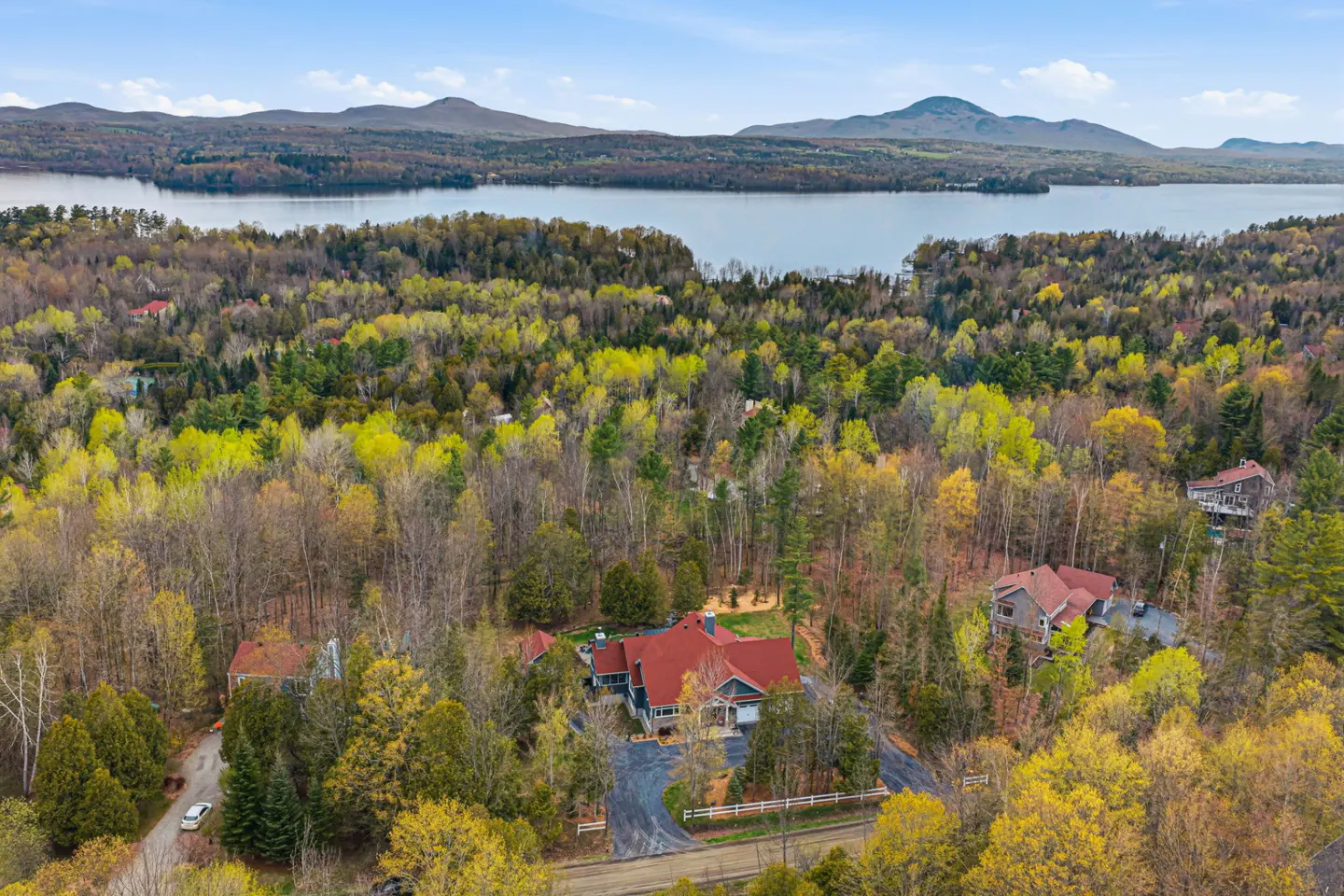Aerial view of a house with a red roof surrounded by trees, with a lake and mountains in the background.