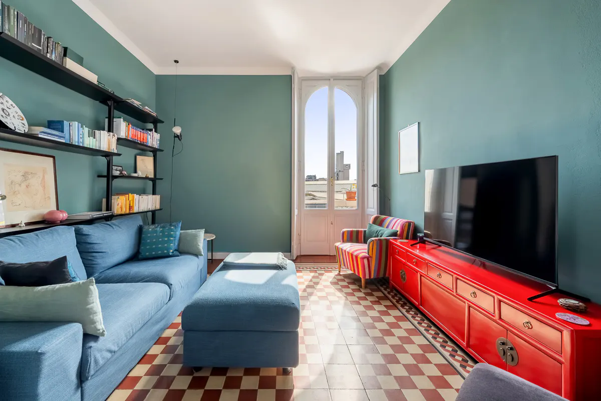 Living room with teal walls, blue sofa, red cabinet with TV, and striped chair. Bookshelves line the wall, and a door leads to a balcony.