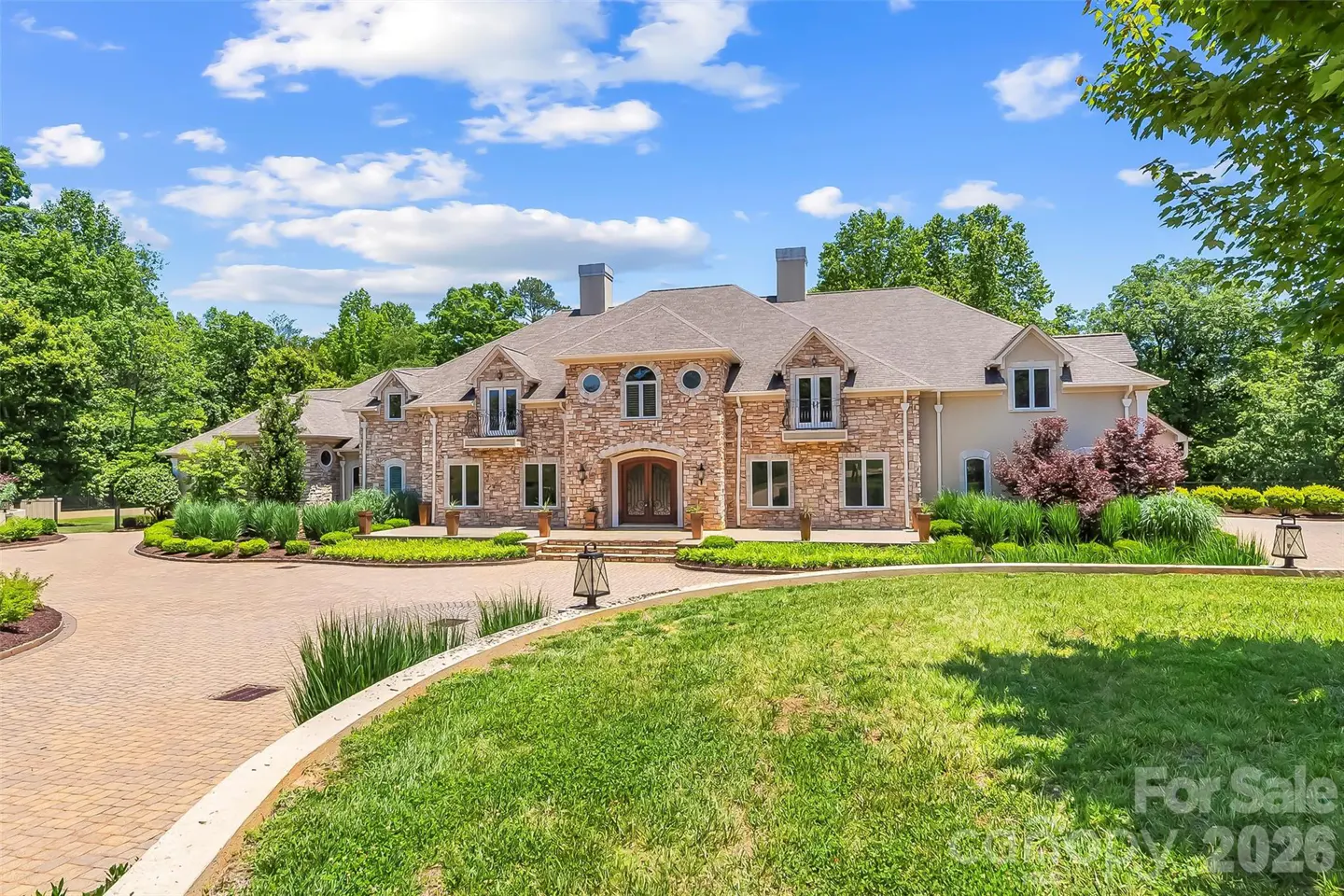 Large stone mansion with a paved circular driveway, lush green lawn, and surrounding trees under a bright blue sky.