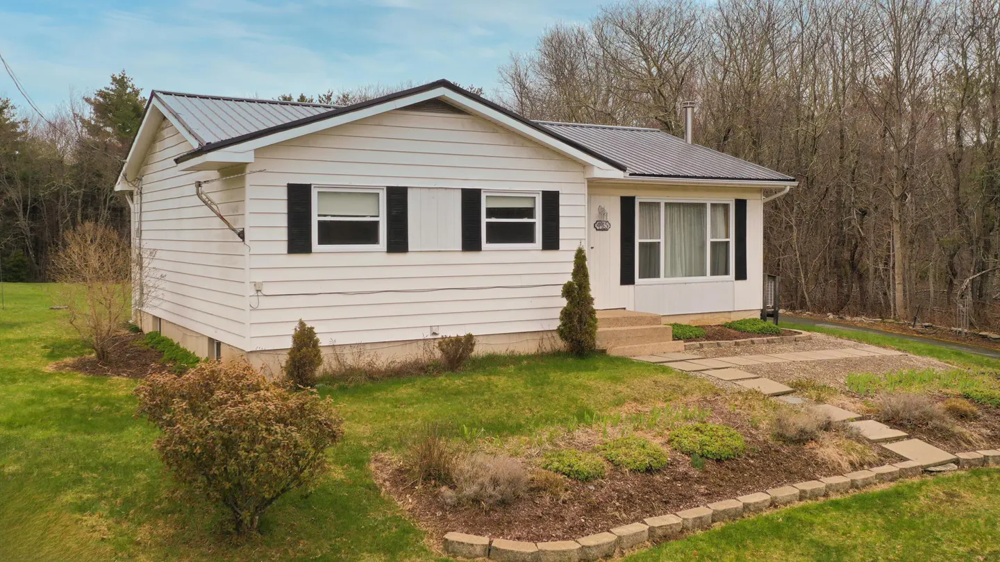 Exterior of a white, one-story house with black shutters and a gray roof, surrounded by a green lawn and trees.