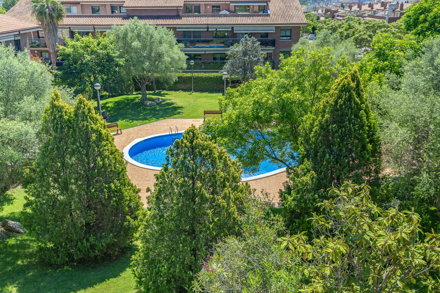 Aerial view of a blue, kidney-shaped pool surrounded by green trees and grass, with a brick building in the background.