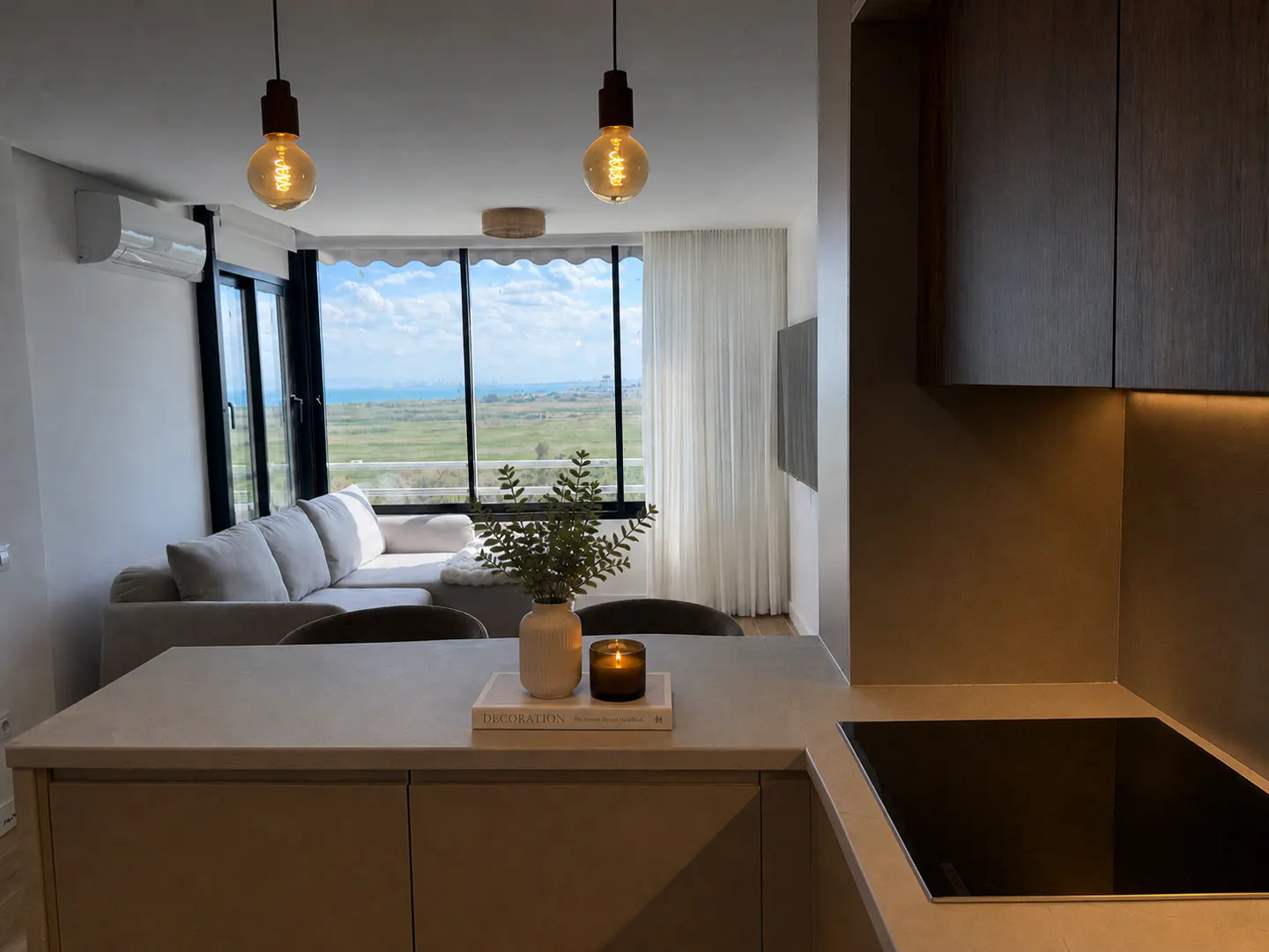A modern apartment interior with a kitchen island, living area, and a scenic view from a large window.
