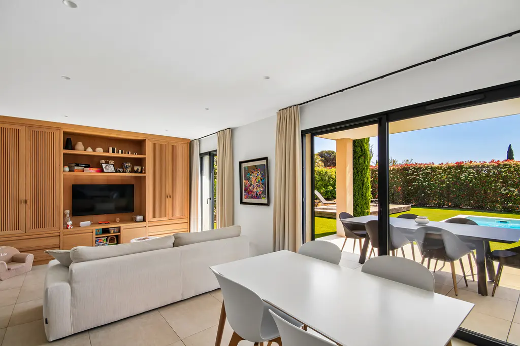 Bright living room with a white sofa, a wooden cabinet, and a white dining table. Outside, a pool and green hedges are visible.