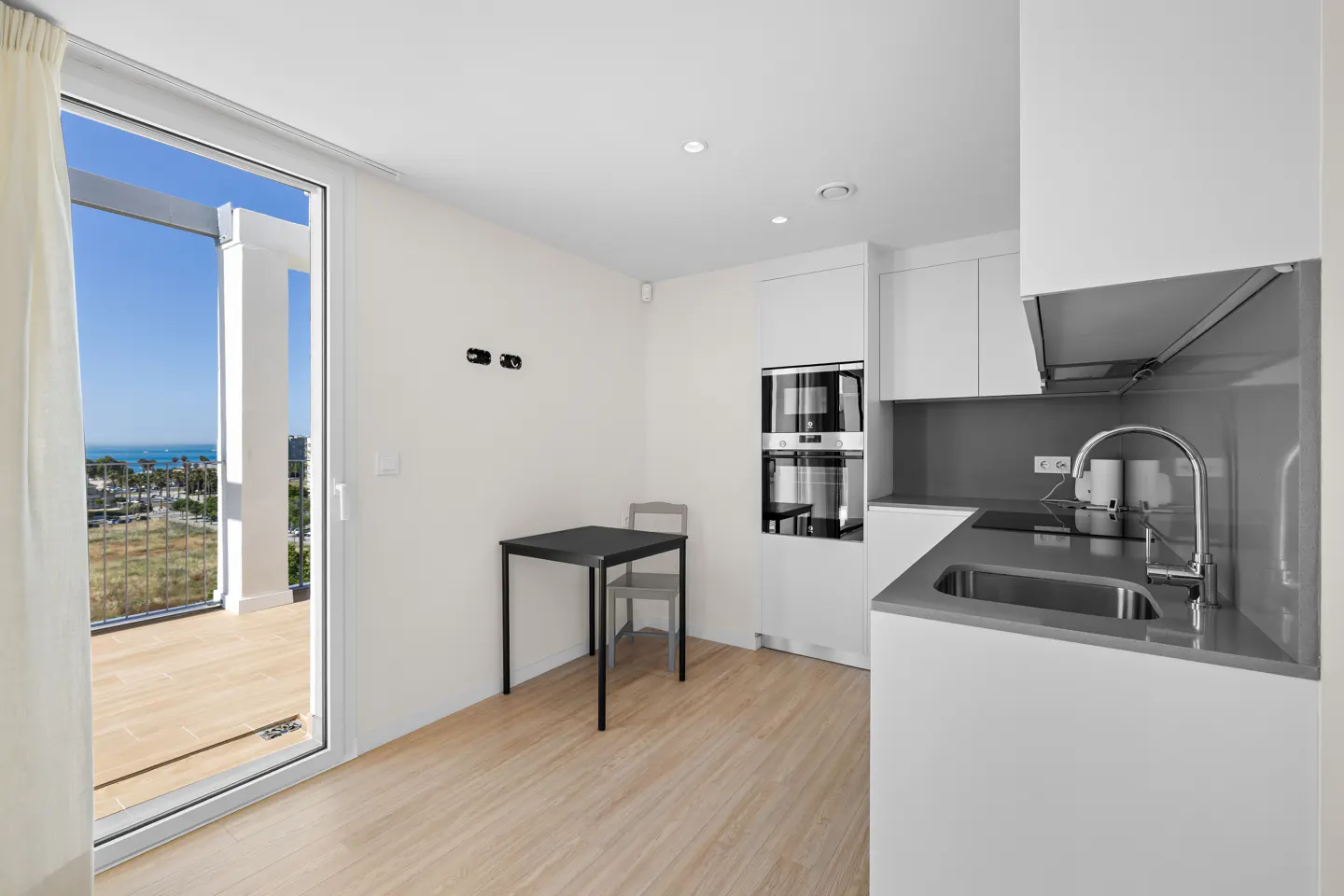 Bright, modern kitchen with white cabinets, gray countertops, and a black table. A sliding glass door opens to a balcony with an ocean view.