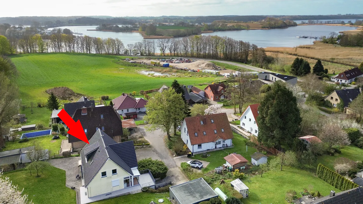 Aerial view of a yellow house with a black roof, indicated by a red arrow, in a green, rural neighborhood near a lake.