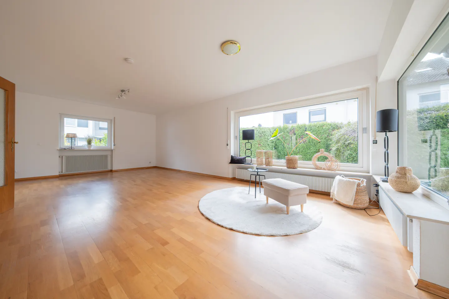 Bright, empty living room with wood floors, white walls, and large windows. A round rug and bench sit in the center.