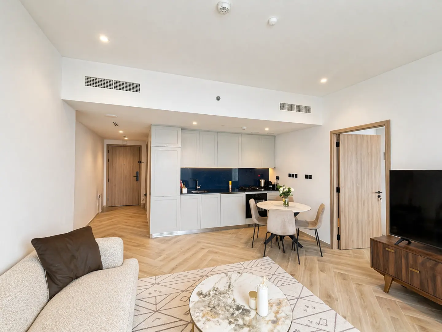 Bright apartment interior with herringbone wood floors, a white sofa, a round marble coffee table, and a kitchen with blue backsplash.