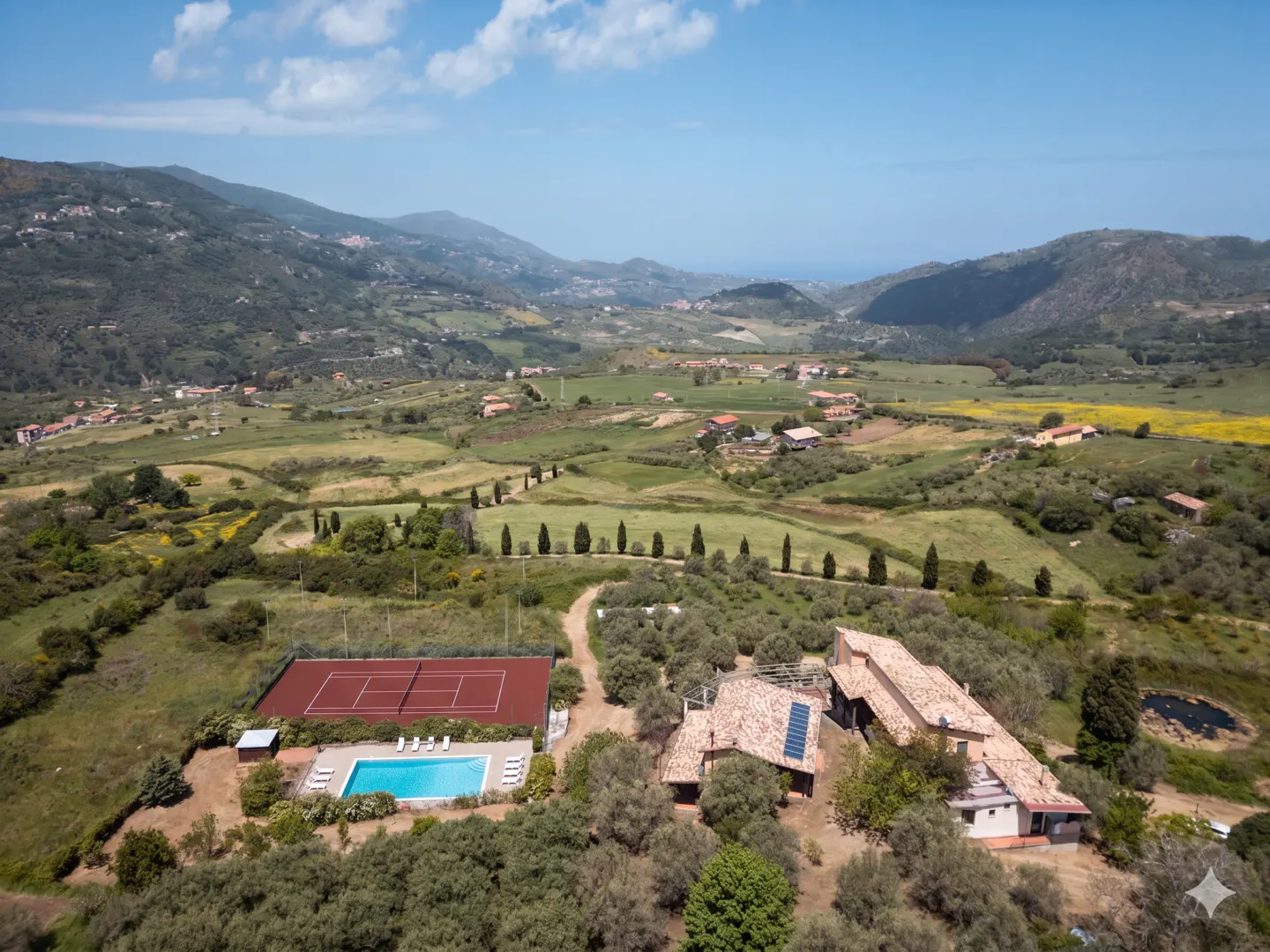 Aerial view of a rural property with a pool, tennis court, and tile-roofed buildings nestled in a green valley with distant mountains.