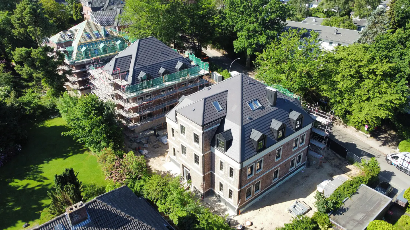 Aerial view of new brick homes with dark gray roofs under construction, surrounded by green trees.