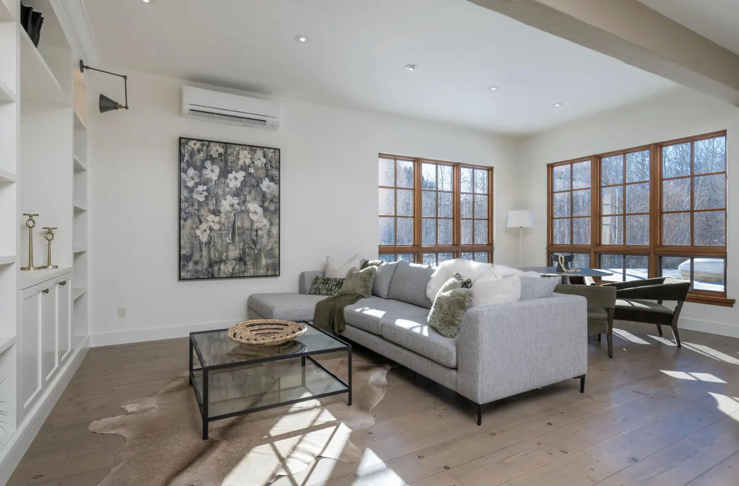 Bright living room with a gray sofa, glass coffee table, and wood floors. Large windows offer a view of trees. A floral painting hangs on the white wall.