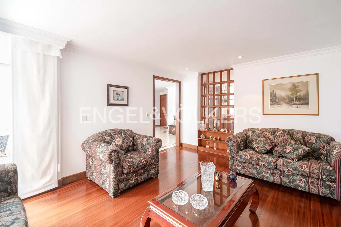 Living room with floral sofas, glass coffee table, hardwood floors, and a wooden room divider leading to a hallway.