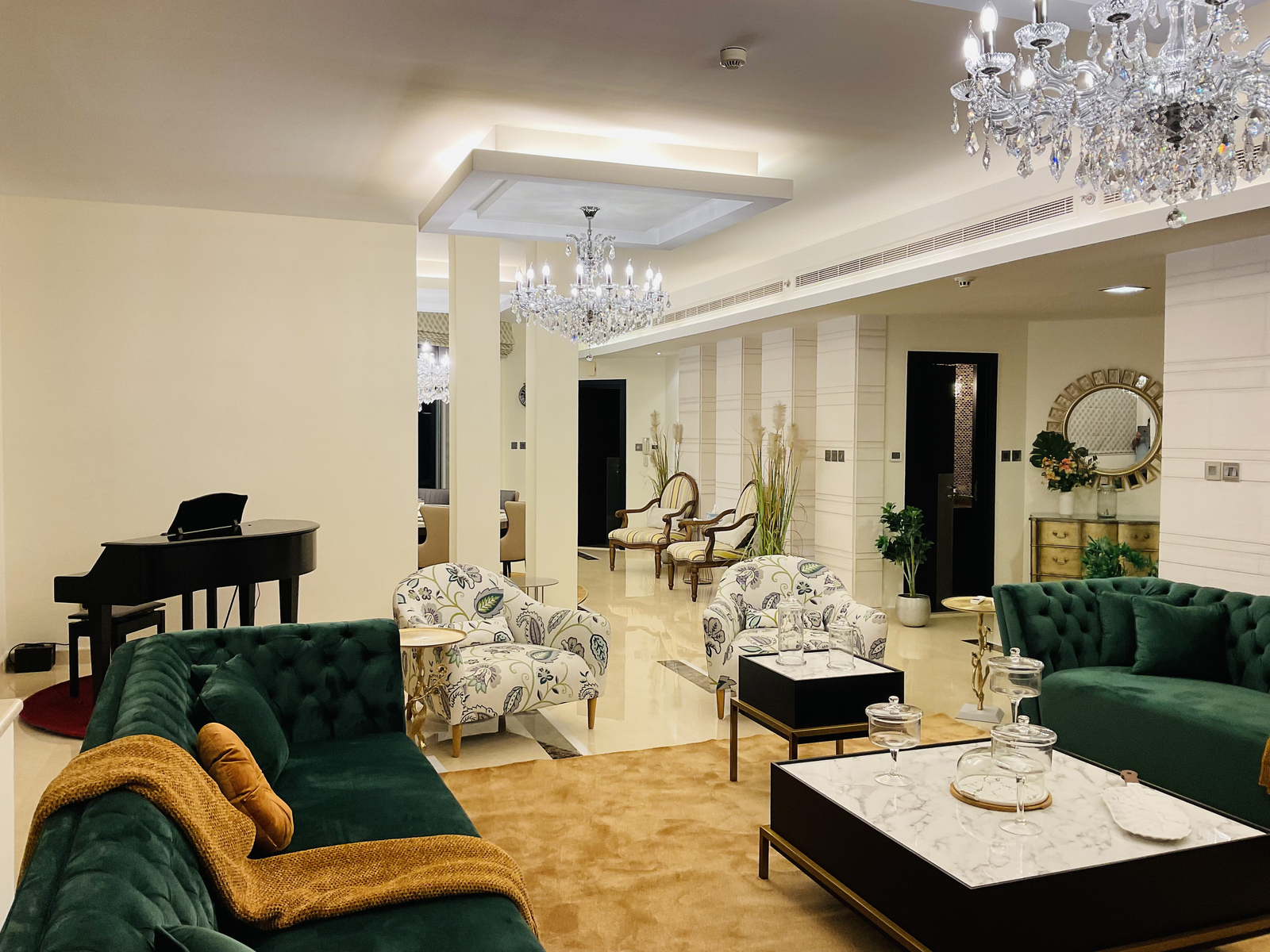 Elegant living room with green velvet sofas, floral armchairs, and a black grand piano. Crystal chandeliers hang from the ceiling.