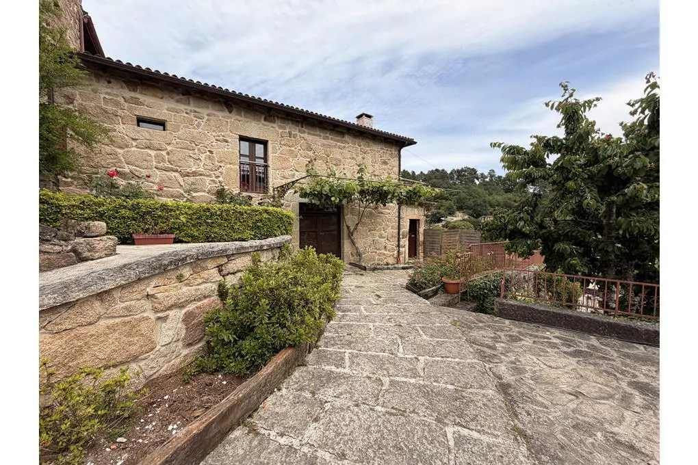 Exterior view of a stone house with a stone path leading to the front door, surrounded by greenery and trees.