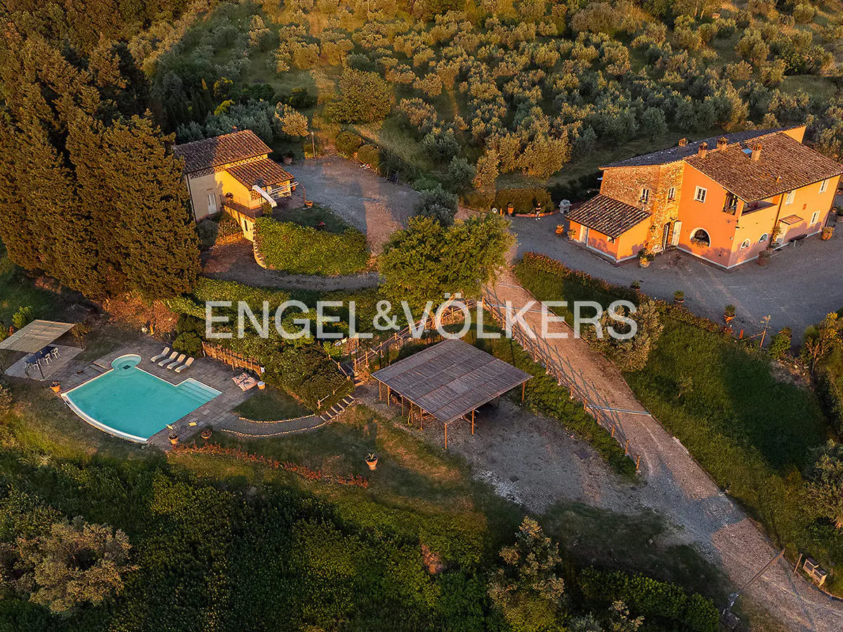 Aerial view of a Tuscan villa with a pool, pergola, and lush green landscape at sunset. Engel & Völkers logo overlay.