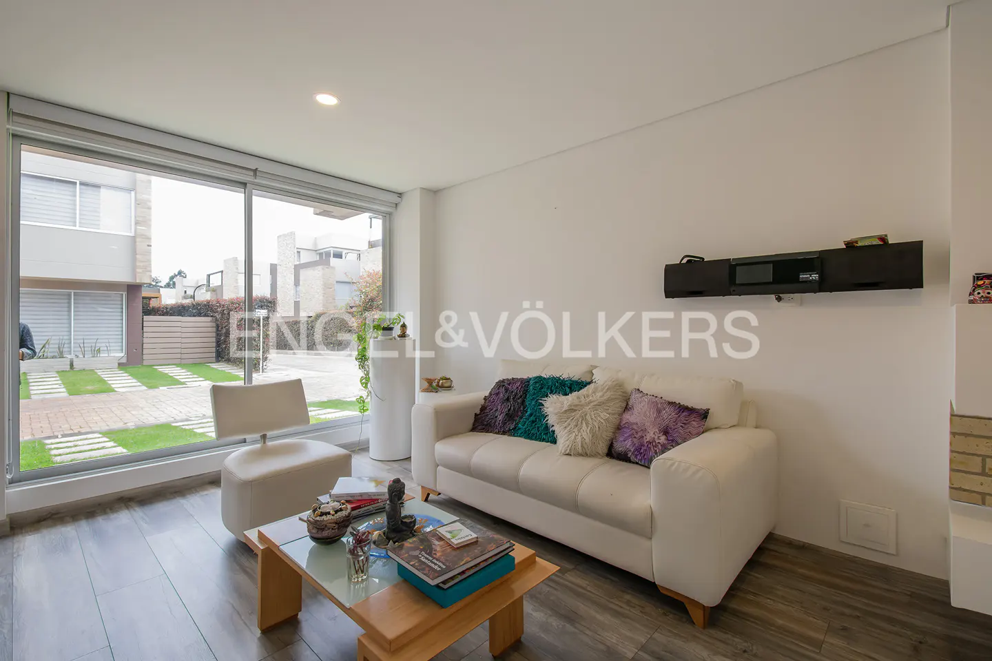 Modern living room with a white sofa, accent pillows, a wooden coffee table, and large windows overlooking a patio.