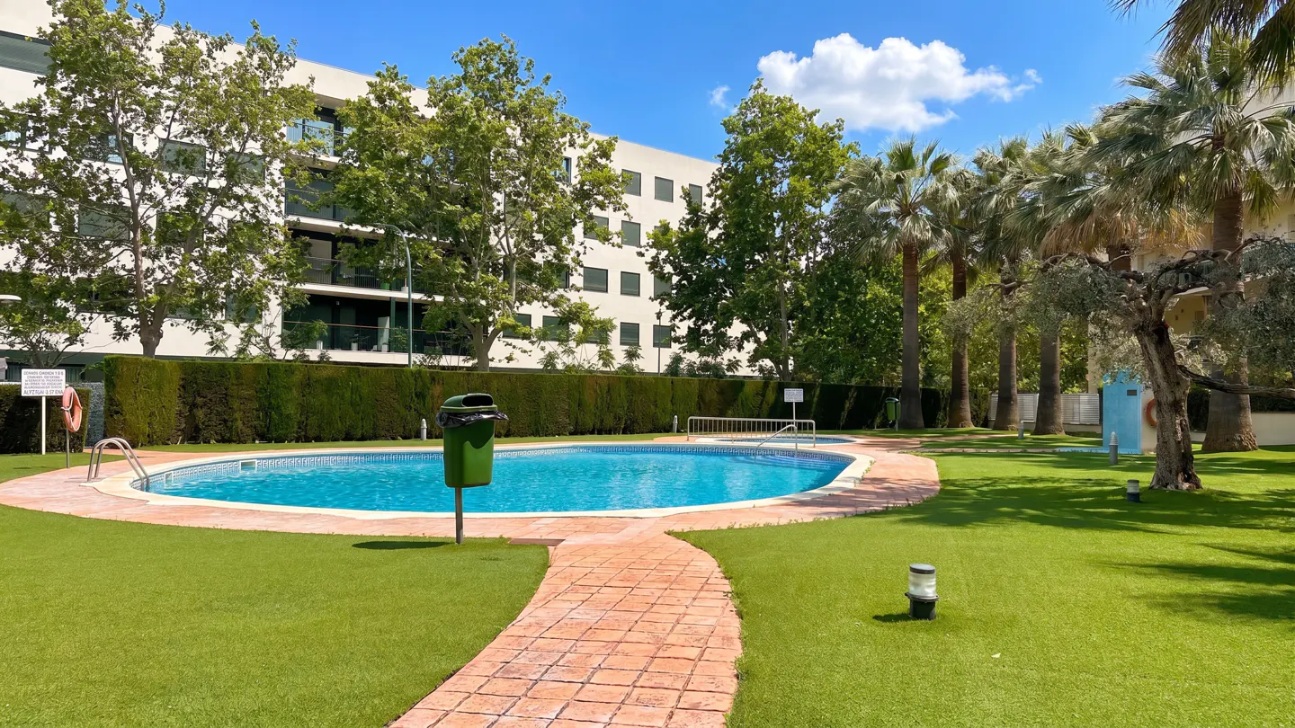 Outdoor pool with turquoise water, surrounded by green grass and a brick path. Tall trees and a building are in the background.