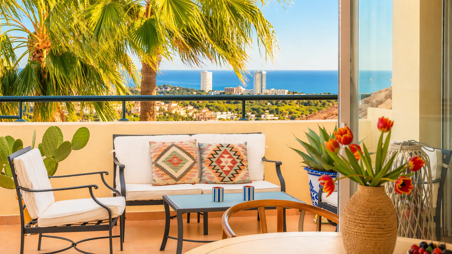 Balcony with outdoor seating, including a white cushioned chair and loveseat with patterned pillows. Ocean view with palm trees and buildings in the distance.