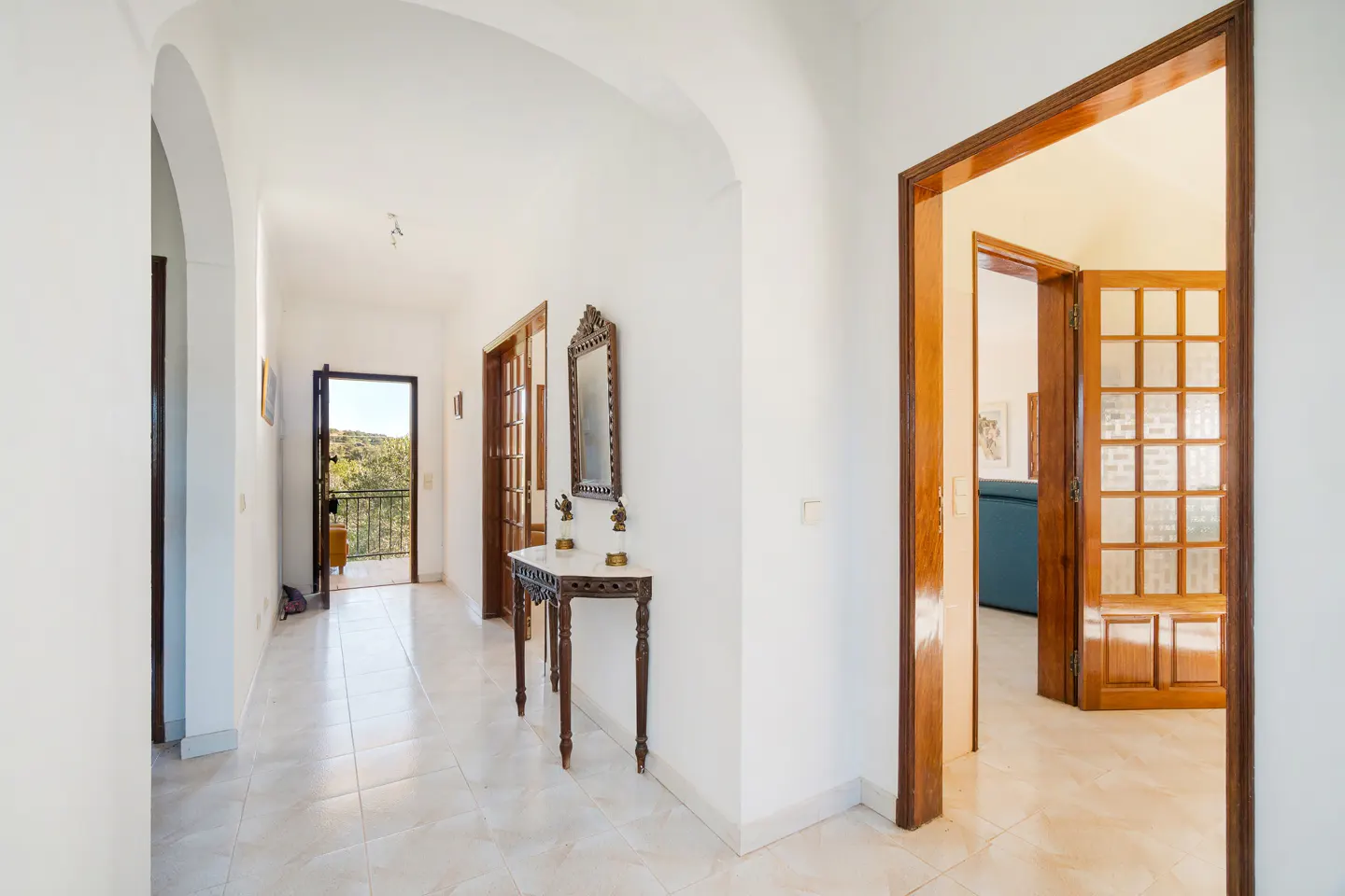 Bright hallway with white walls, arched doorways, and tiled floor. A dark wood console table with a mirror stands against the wall. An open door leads to a balcony.