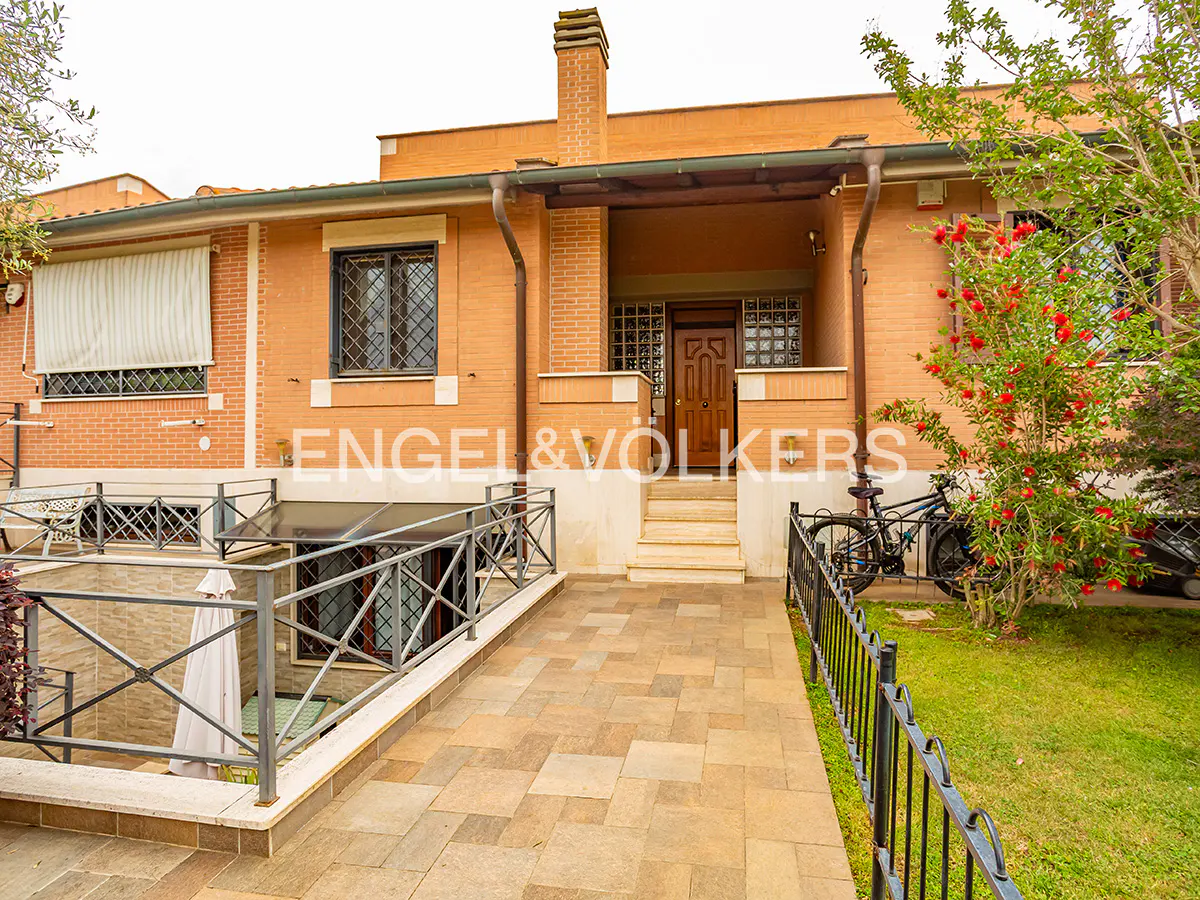 Exterior view of a two-story brick house with a stone walkway, black metal railings, and a brown front door. A bicycle is parked near a tree with red flowers.