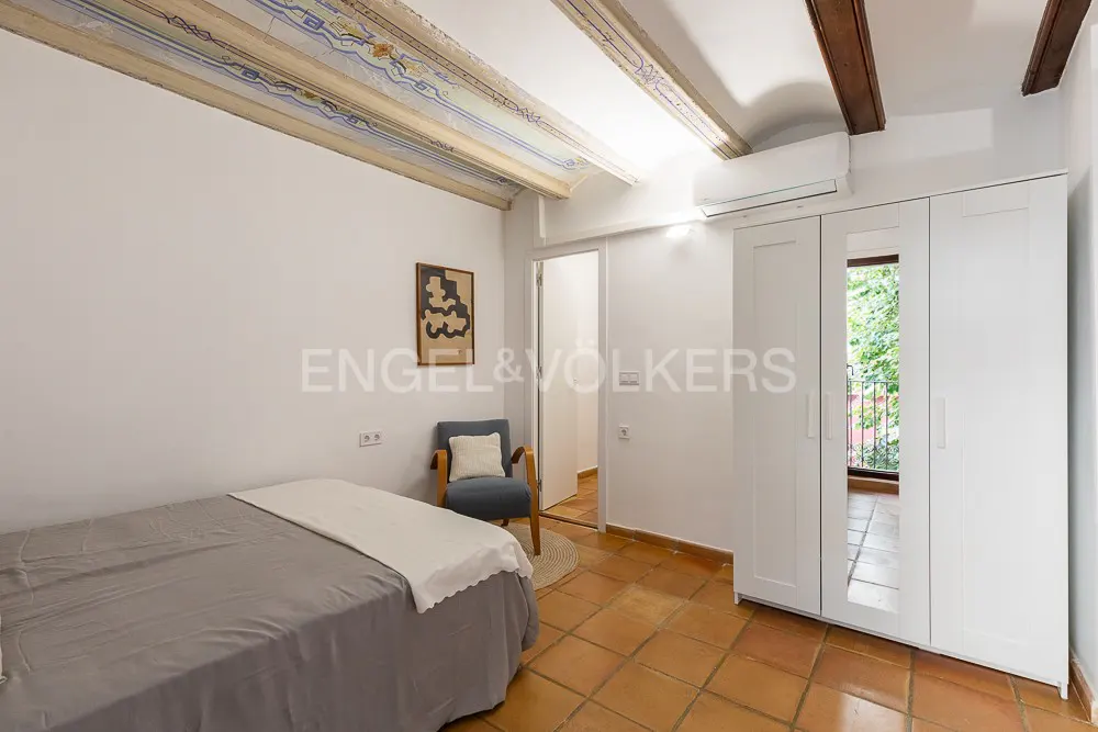 Bedroom with terracotta tile floor, white walls, and painted ceiling beams. A grey bed, blue chair, and white wardrobe furnish the space.