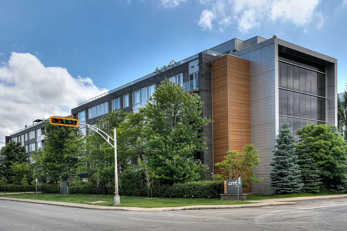 Exterior view of a modern, multi-story apartment building with mixed materials, green trees, and a traffic light.