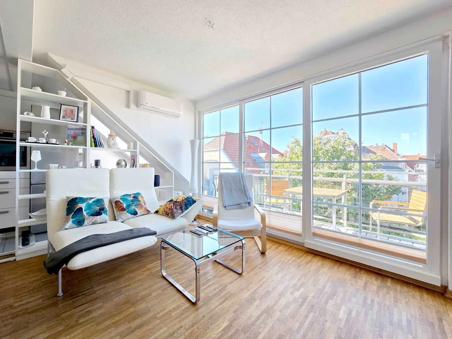 Bright living room with white sofa, glass table, and large windows leading to a balcony with outdoor furniture.